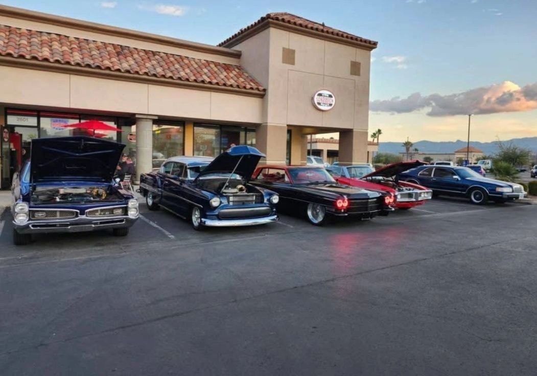 The Sandwich Spot Henderson storefront at dusk, with its red oval sign and a row of classic cars parked out front